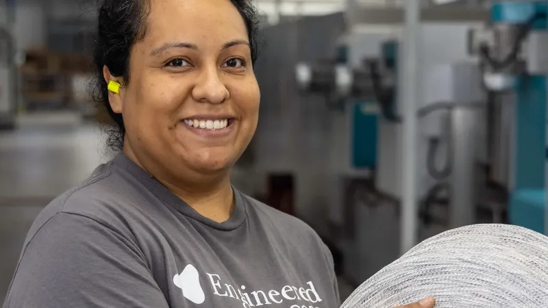 A smiling person holding a roll of carpet material in a flooring manufacturing facility.