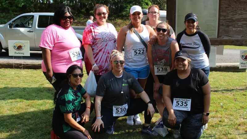 A group of nine people posing outdoors after a trail run event, wearing athletic attire with numbered race bibs.