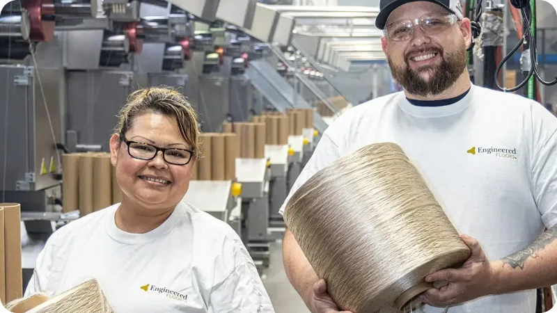 Two employees wearing Engineered Floors shirts hold spools of carpet fiber in a manufacturing facility.